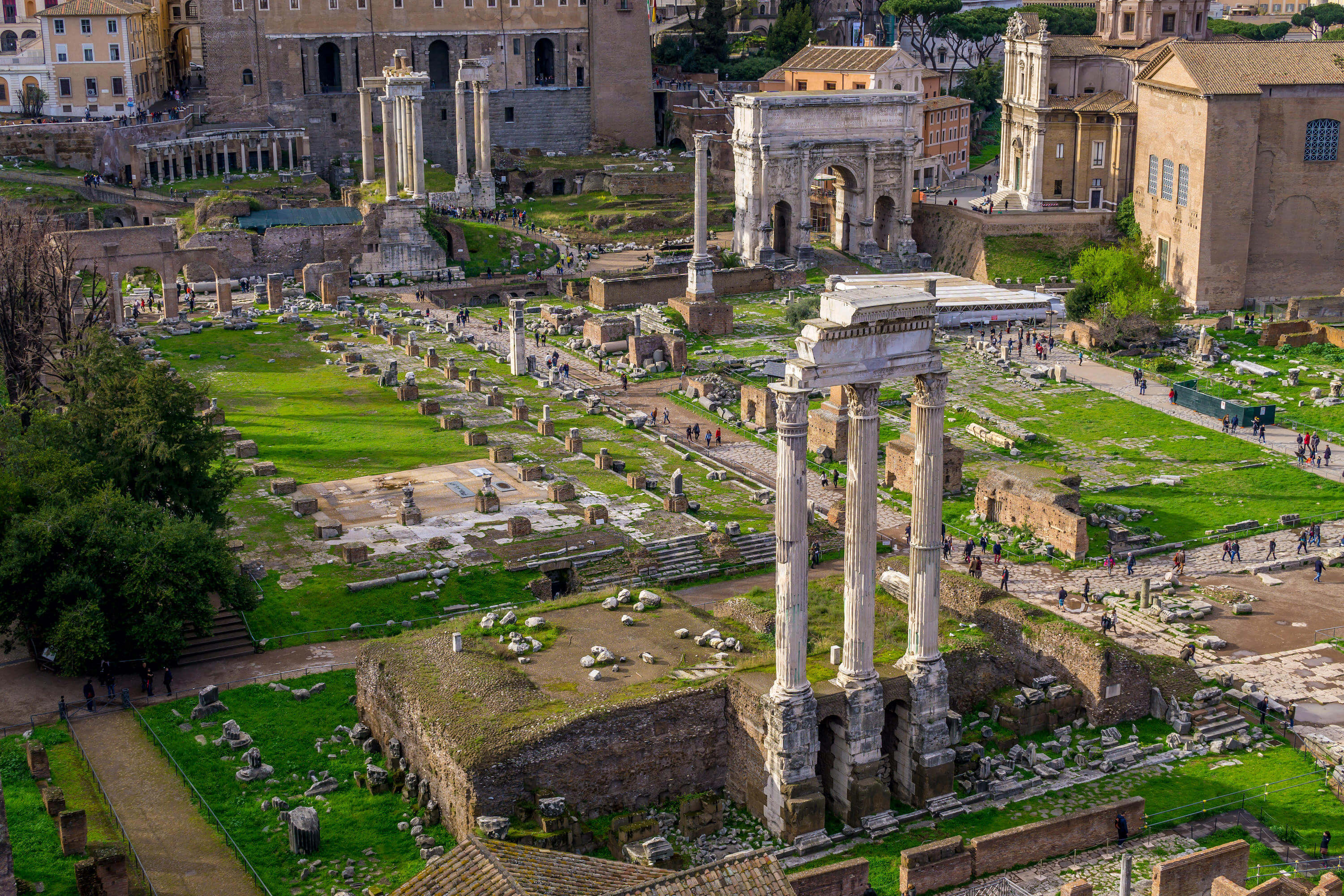 A photo taken from a higher vantage pint looking down on the remains of the Roman forum. Some columns remain standing and on the right is a stone arch.