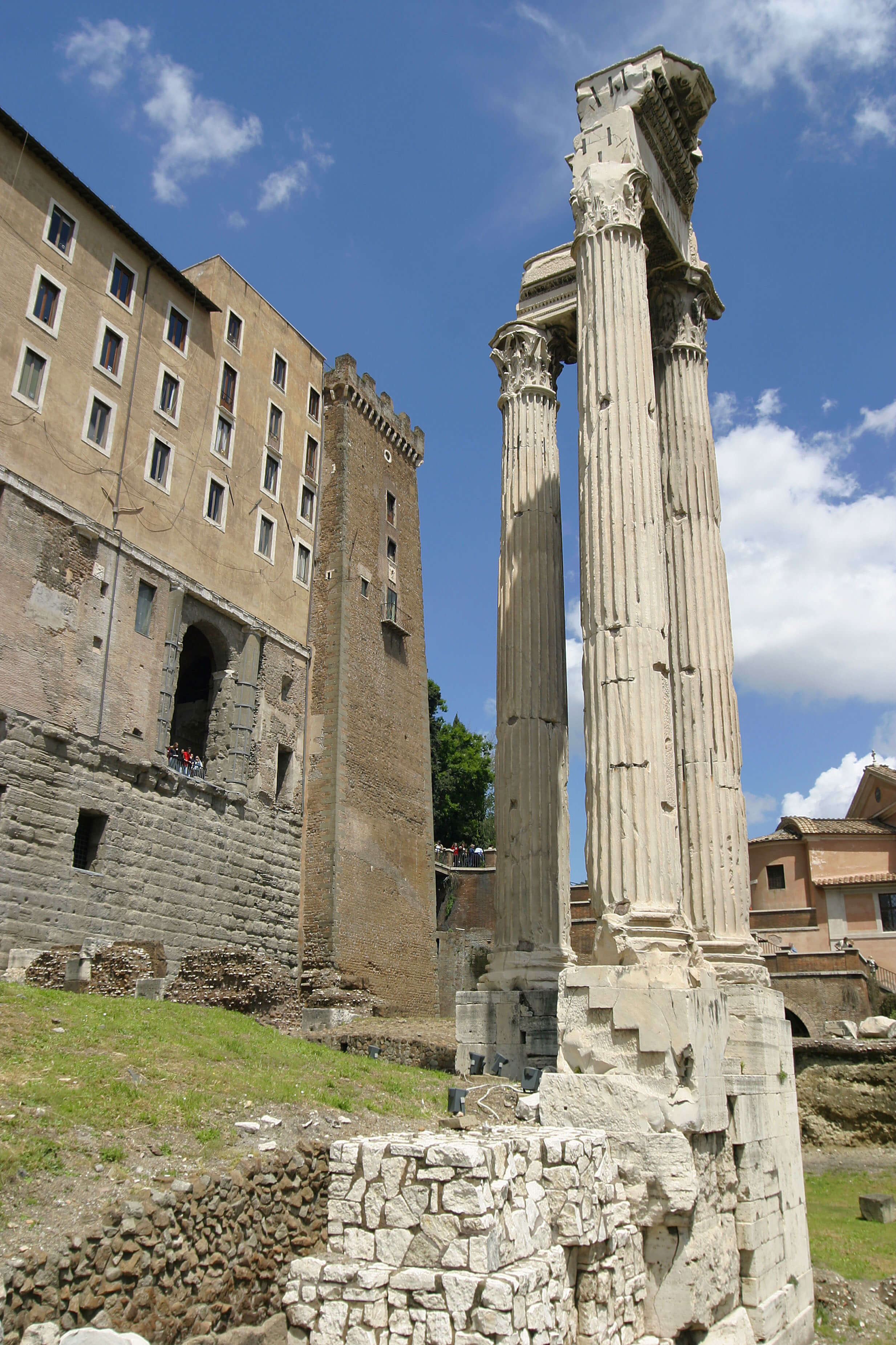 Three Roman columns standing on a raised platform. The other remains of the temple are no longer standing. On the left is a modern building with stairs up to the front.
