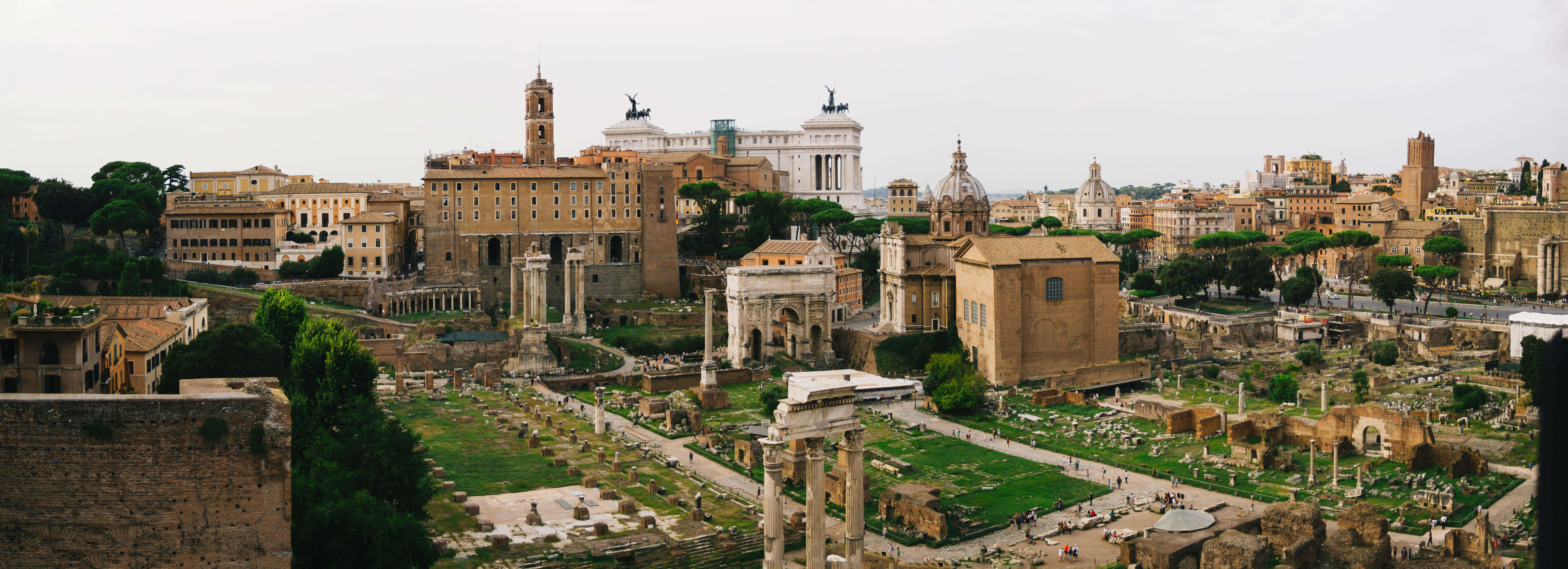 A panoramic view of the remains of the Roman Forum. A few columns stand at their original height, but most are stumps. One arch remains standing. In the background is the modern city of Rome and there are domes and spires.