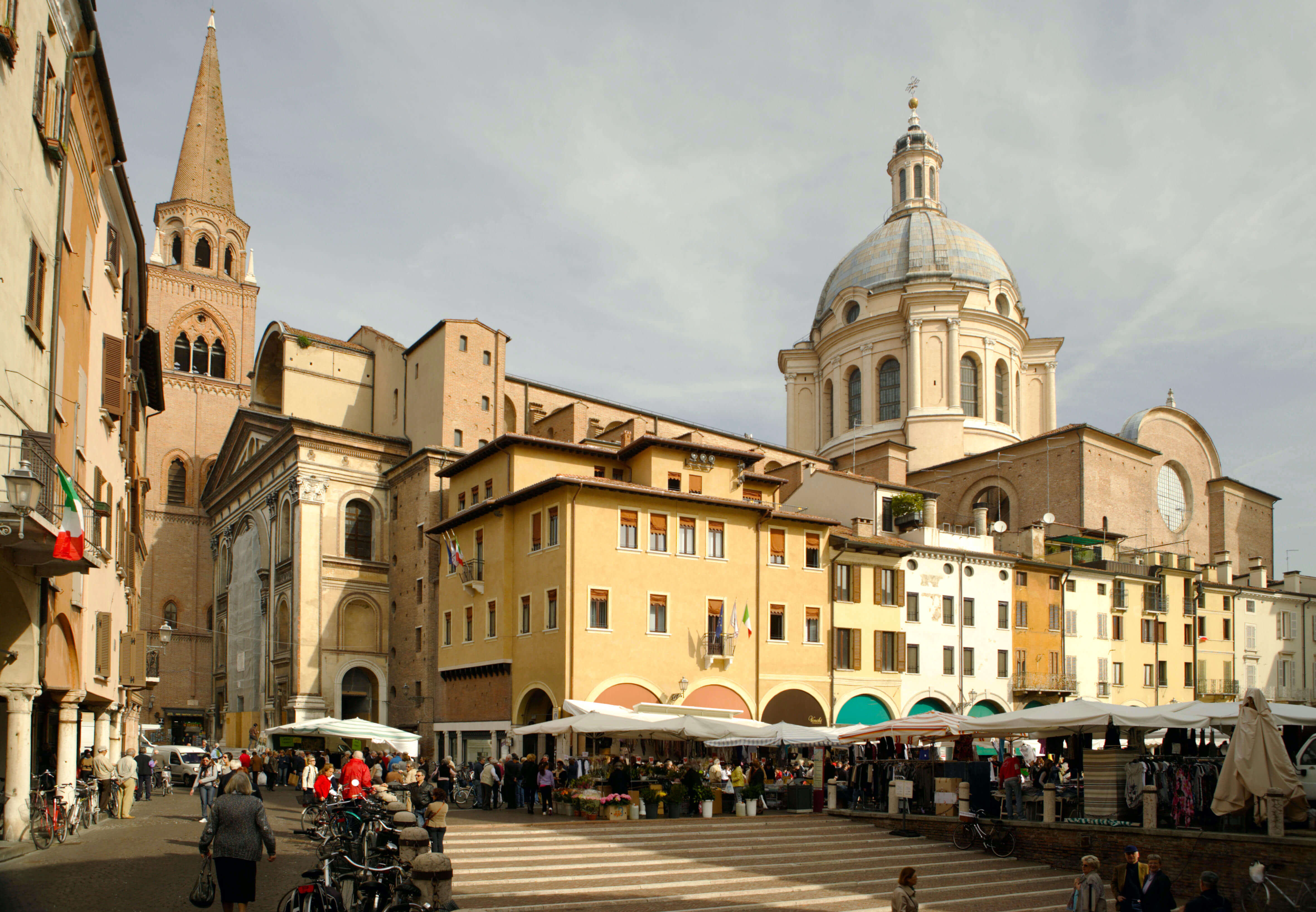 A modern townsquare. It is busy with activity and there are people shopping, chatting, and walking around at the market stalls. There is a church spire and the dome of a second church. Some buildings have Italian flags hanign from balconies.