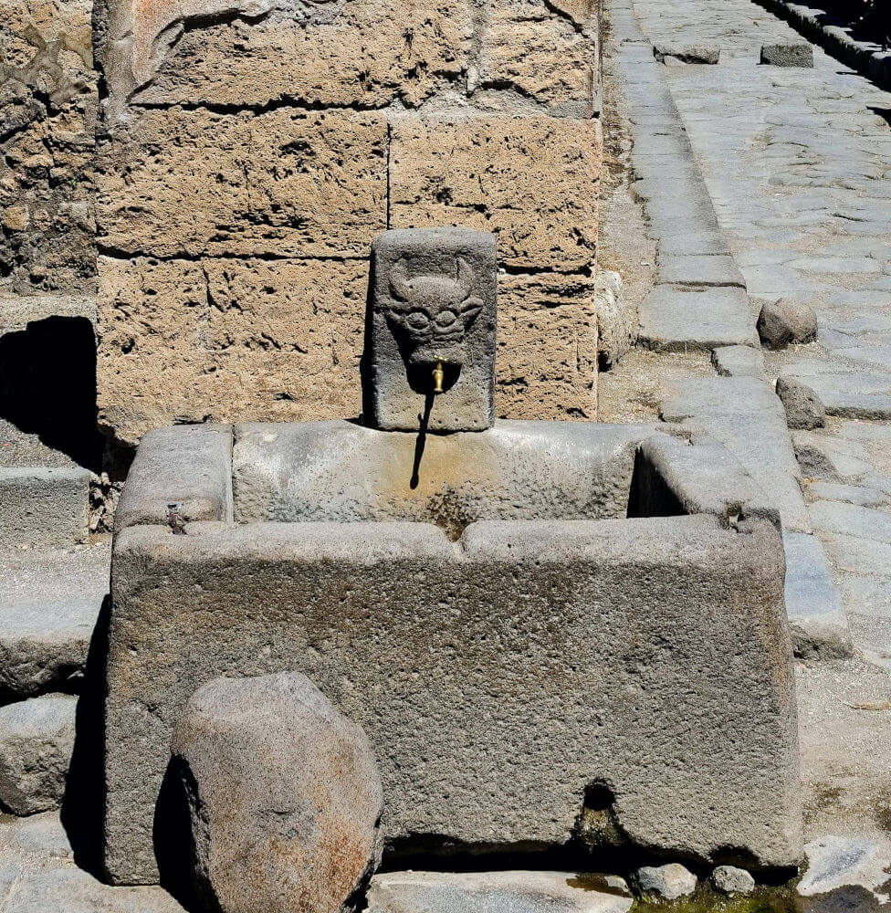 A stone water fountain on a street corner. The tap of the fountain is coming from the mouth of a carved bull head.
