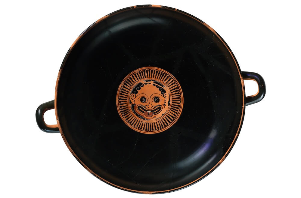 A round drinking cup with two handles. It is black and in the centre there is a female head in a patterned circle. The face has wide eyes and a large mouth.