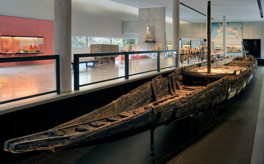 An image of an ancient wooden barge in a museum, in remarkably good condition. It is surrounded by light-coloured amphorae.