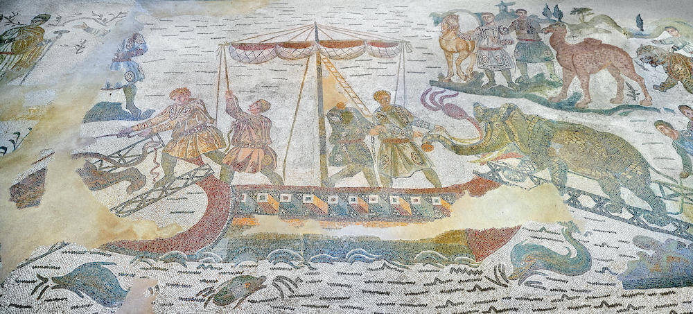 A mosaic depicting wild beasts being loaded onto a ship. On the right an elephant is being led up the gang plank. Behing there is a camel. In the sea dolphins swim.