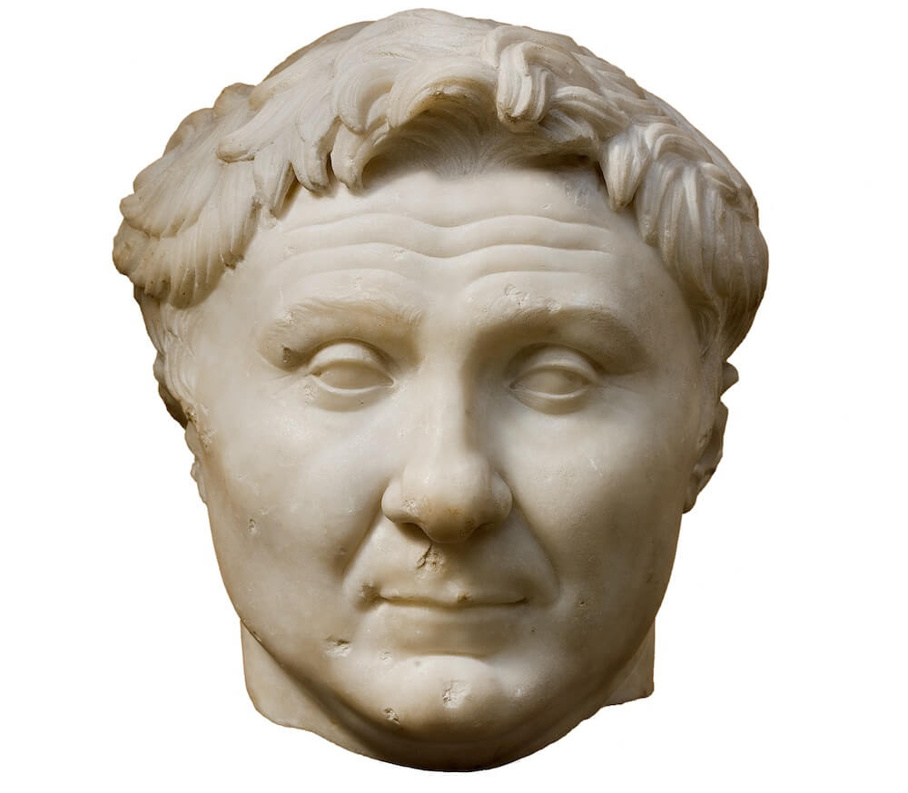 A stone head of a man with a round face and a flick of hair on his forehead.