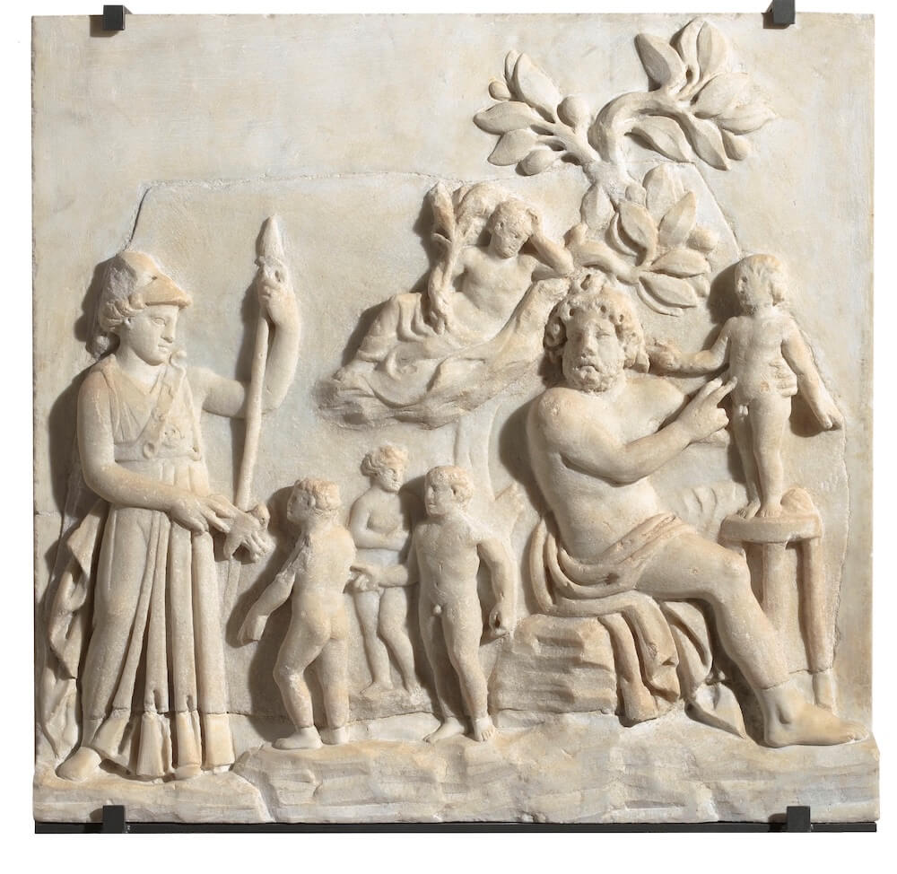 A stone relief showing a man sitting on a chair with a much smaller figure on a stool next to him. On the left is a goddess wearing a helmet and holding a spear.