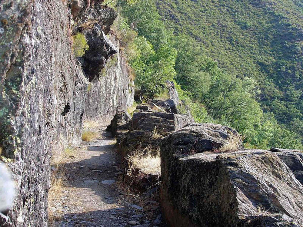 A photo of a path cut into a mountain side. The side is very rough and ahead here are many trees.