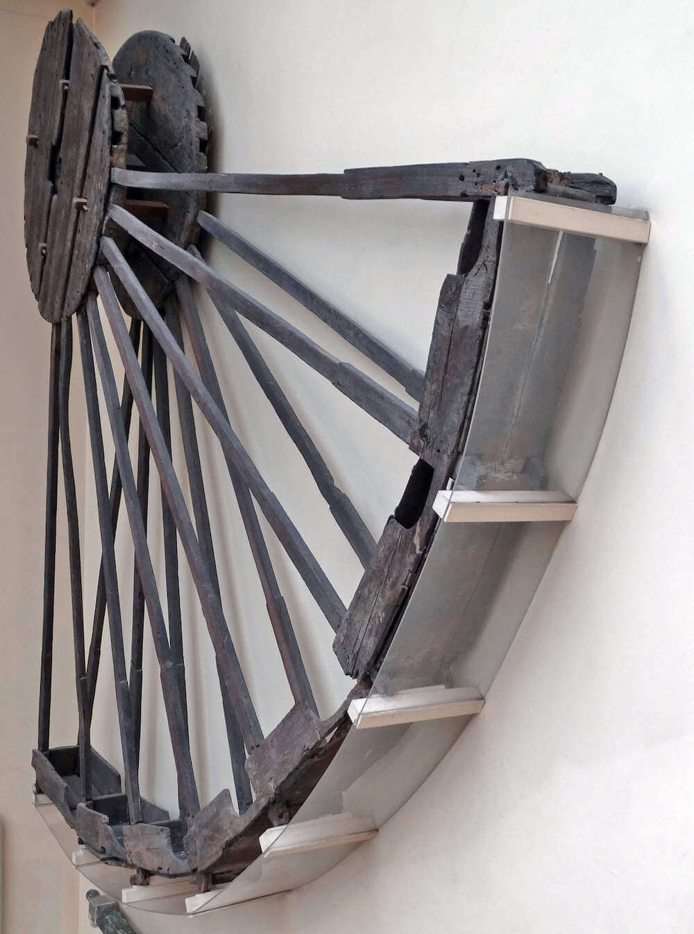 A photo of a wooden wheel mounted on a wall. Only about a quarter of it survives and it is held in a protective glass casing.
