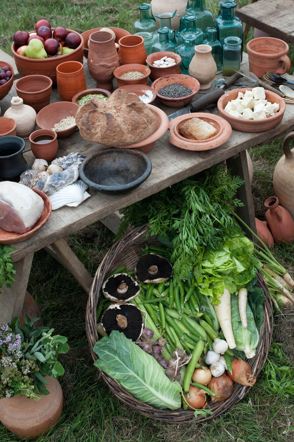 A photo of a table of raw foods and ingredients. In a basket under the table are mushrooms, parsnips, onions, beans, and cabbages. On the table is a loaf of bread, oyster shells, as well as bowls and jars of various ingredients. 