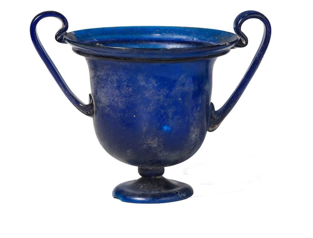 A cup with two arching, thin handles. It is made of blue glass and is full intact.