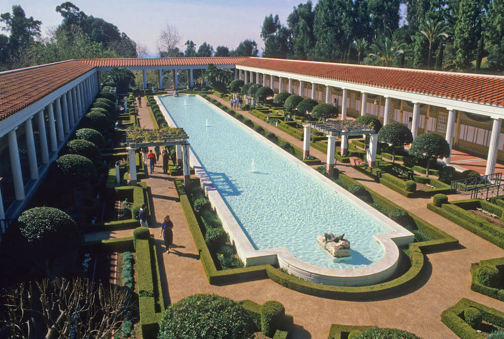 A photo of a garden surrounded by a roofed portico. In the middle of the garden is a long pool and the shrubs and trees are all trimmed in an orderly fashion.