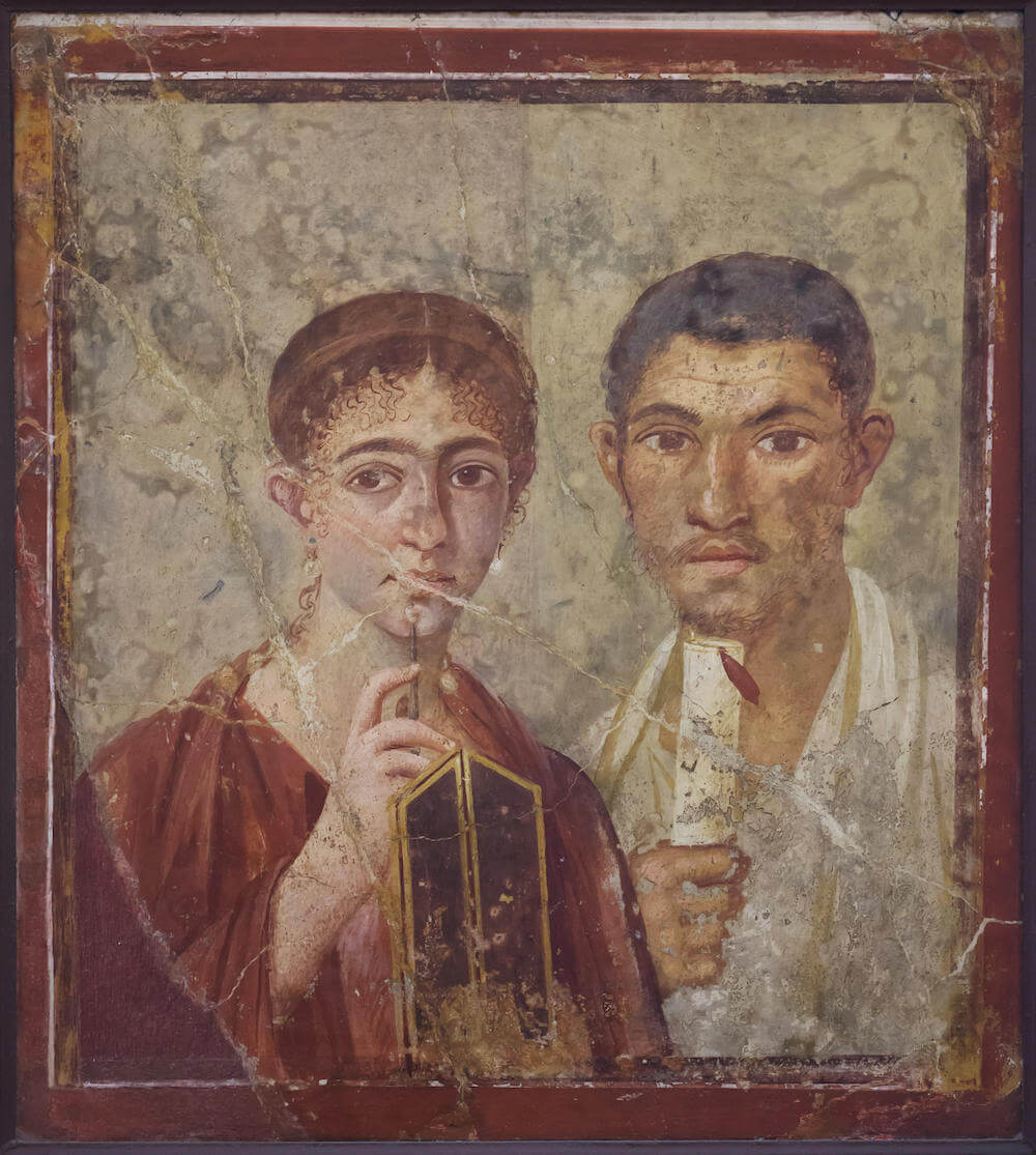 A wall painting showing a man and a woman facing forward. The woman is holding a wax writing tablet and a stylus. The man is holding a scroll and is wearing a white toga.