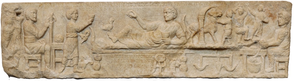 A long carved stone sarcophagus. It shows a man reclining on a bed holding a cup. On his left men are leading an animal through an arch and a man is spinning. On the left a woman offer a cup and a man sits on a chair.