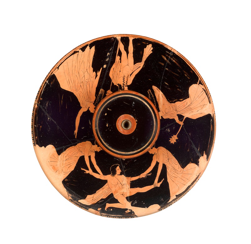 A wide, flat bowl with a black background and figures picked out in red. In the centre a man kneels with his arms and legs stretched out. On either side of him a woman holds his limbs and pulls.