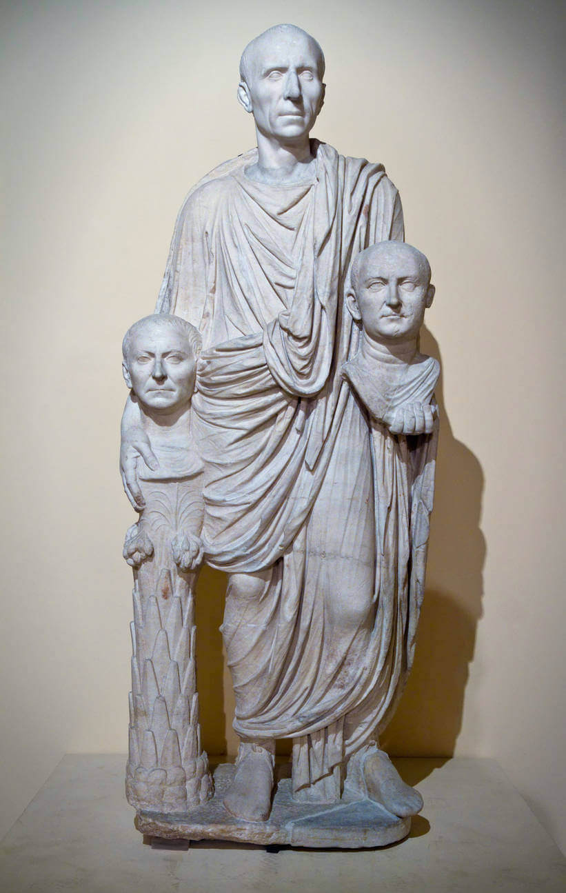 A statue of a man wearing a toga. In each hand he holds a bust of another man, one is resting on a plinth.