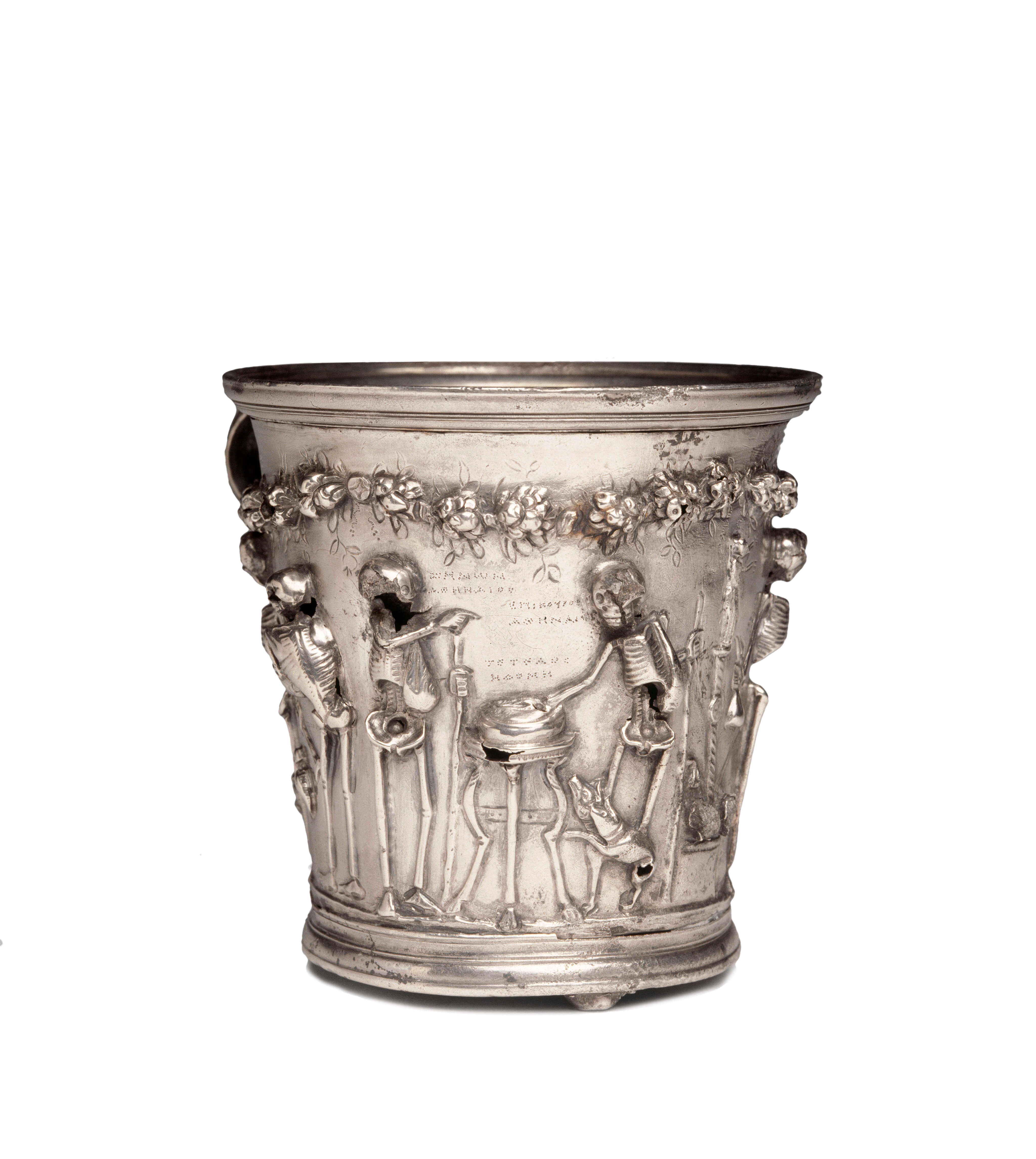 A silver cup decorated with images of skeletons. On this side two skeletons face each other across a small table. By the bottom right of the table is a piglet. Above their heads hangs a garland of flowers.