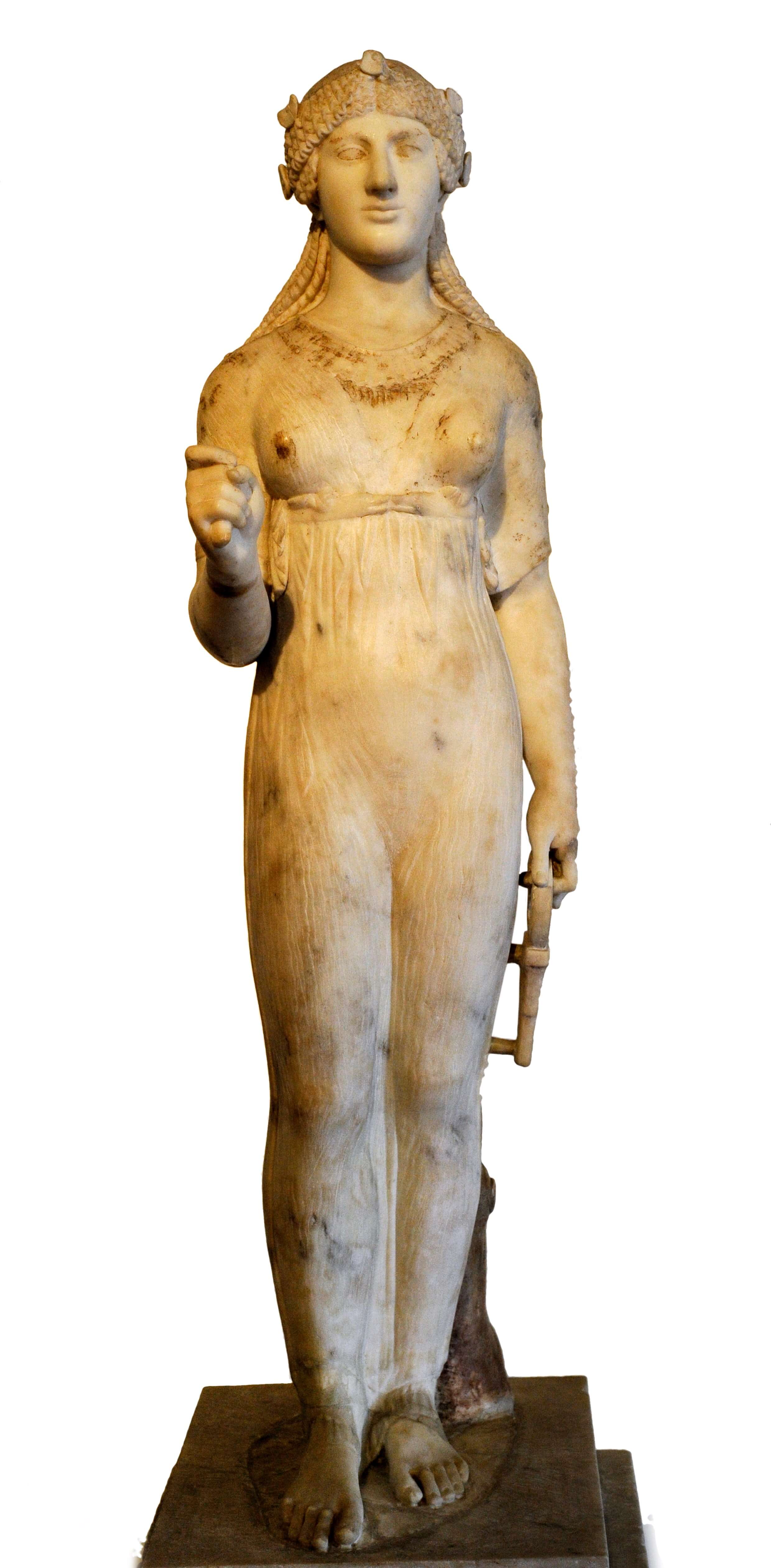 A statue of a woman. She has braided hair tied back from her face. She is wearing a light dress through which you can see the curves of her body. Her right hand is raised and her left hangs by her side holding an object.