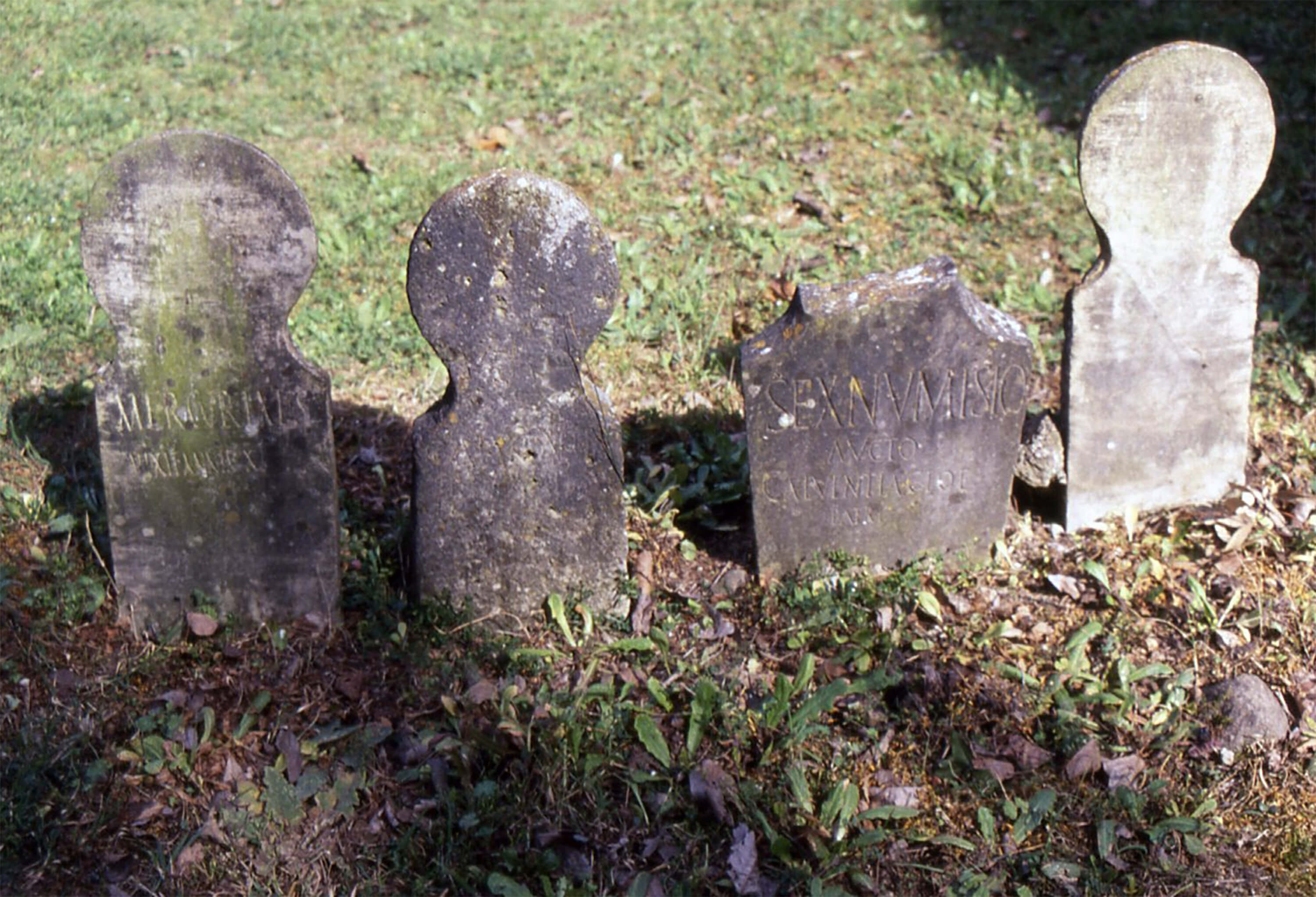 Four small rectangular slabs of stone with a circular head at the top. On it is carved words in capital letters. They are half buried in the earth adn stand upright.