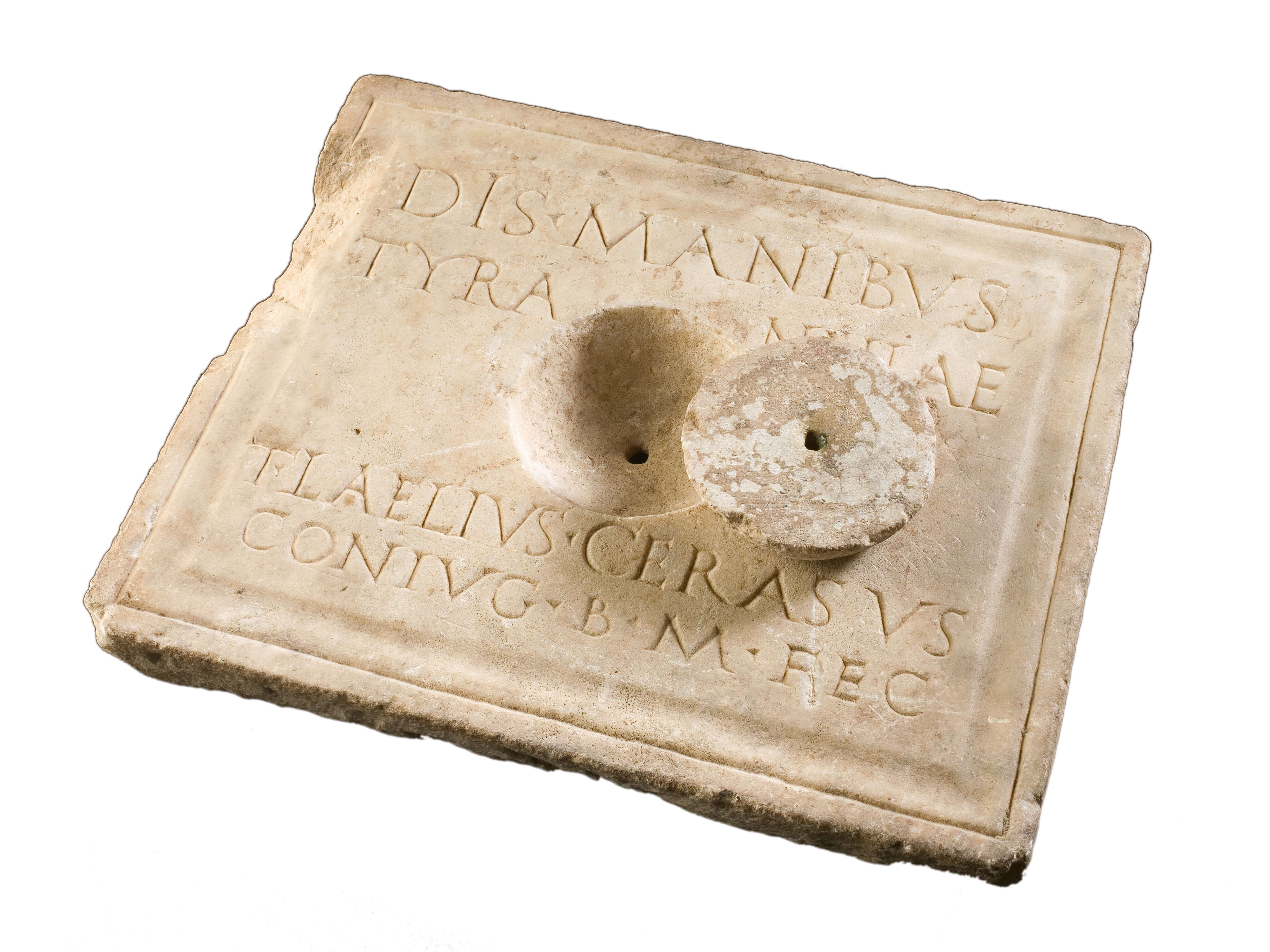 A stone slab with capital letters neatly marked on it. In the centre is a circlar hollow with a hole at the bottom. The is also a stone lid to cover this hollow.