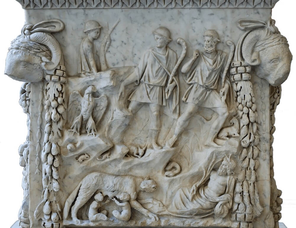 One side of a stone altar. It is intricately carved. At the bottom left a wolf suckles two young children in a cave. On the bottom right an old man with a beard reclines. Above, an eagle sits on the left and two young men wearing caps stand. On the top left a shepherd boy sits with a staff. On either side of the image are two ram heads and garlands of vines.
