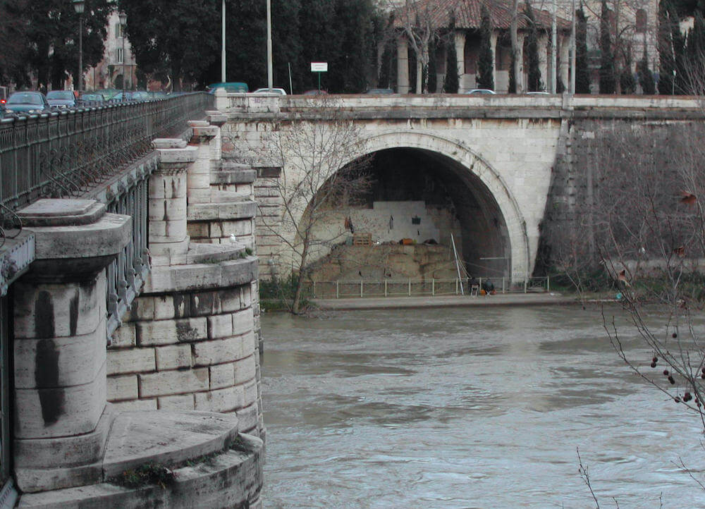An arched tunnel opening onto the a river. On the left a large bridge crosses the river. The tunnel has been bricked up.
