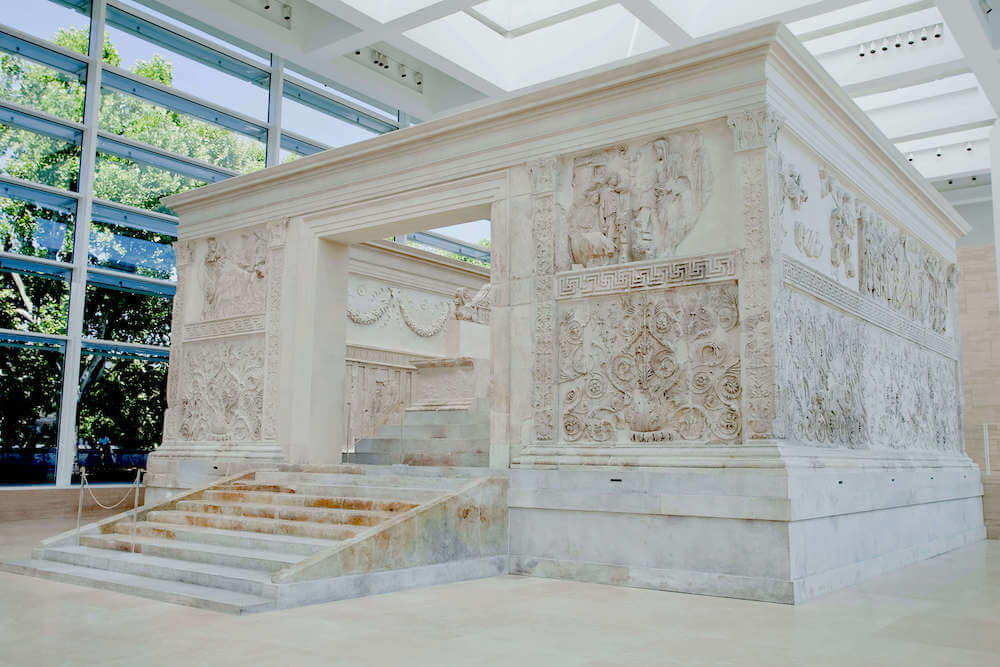 A large white stone monument with carvings on the walls. One side has an opening and stairrs goign up to the inside.