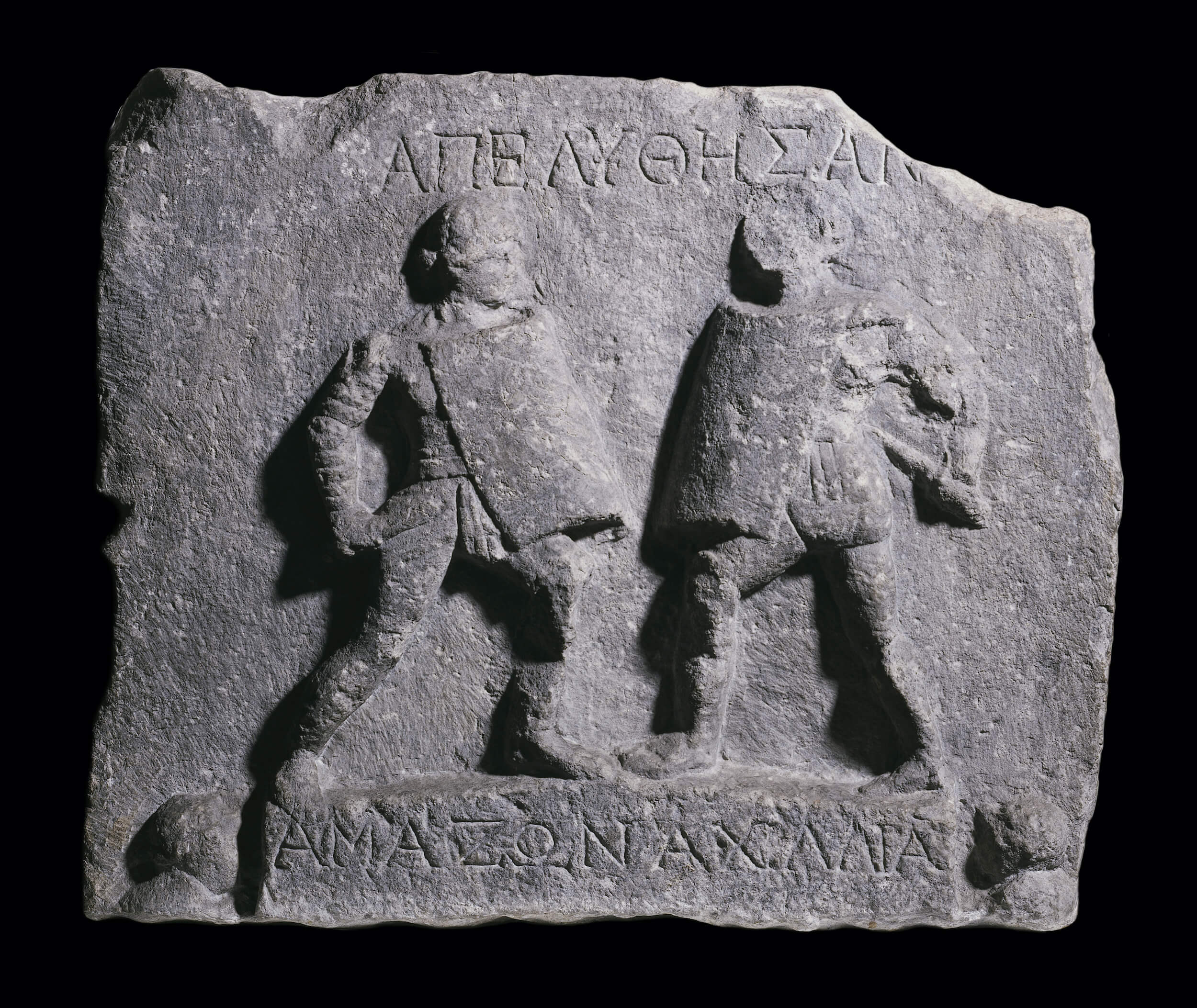 A stone carving showing two gladiators standing on a small stage fighting. The are greek letters carved above their heads and below their feet. On either side of the stage are the heads of spectators.