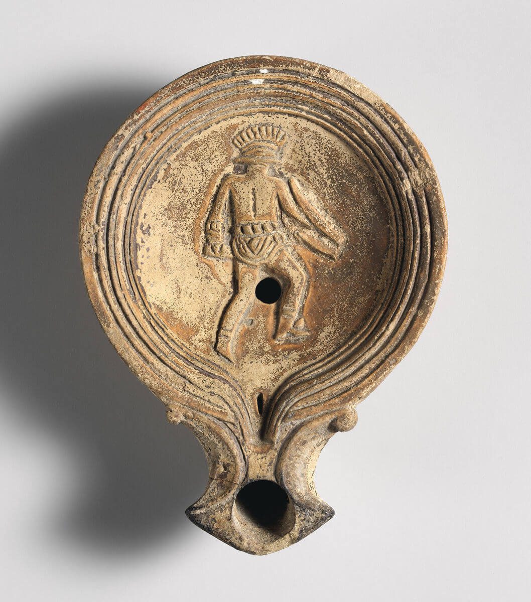 A clay oil lamp. On the top is carved a gladiator with a crested helmet.
