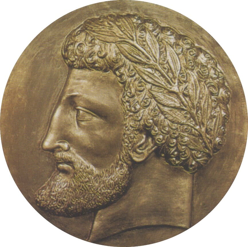 A dark metal coin with the side profile of a man with a pointed beard and a band tied around his head.
