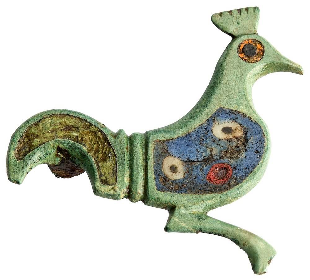 A green brooch in the shape of a cockerels seen from the side with decorated body and a bright eye.
