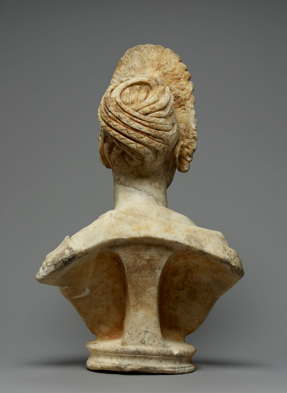 A stone bust of a woman seen from behind. Her hair is arranged in a complicated style wrapped around at the back.
