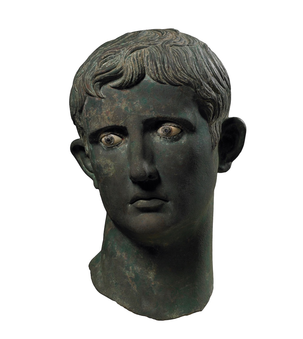 A metal head of a young man. It has become dark and green-coloured over time. The hair is arranged in locks on the forehead and the whites of the eyes stand out brightly.