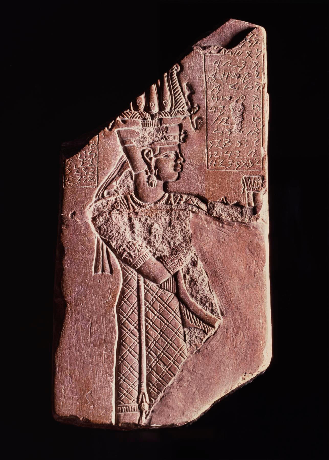 A fragment of red-coloured stone with an engraving of a woman standing side on with an elaborate headdress. To the right of head head is a column of script.