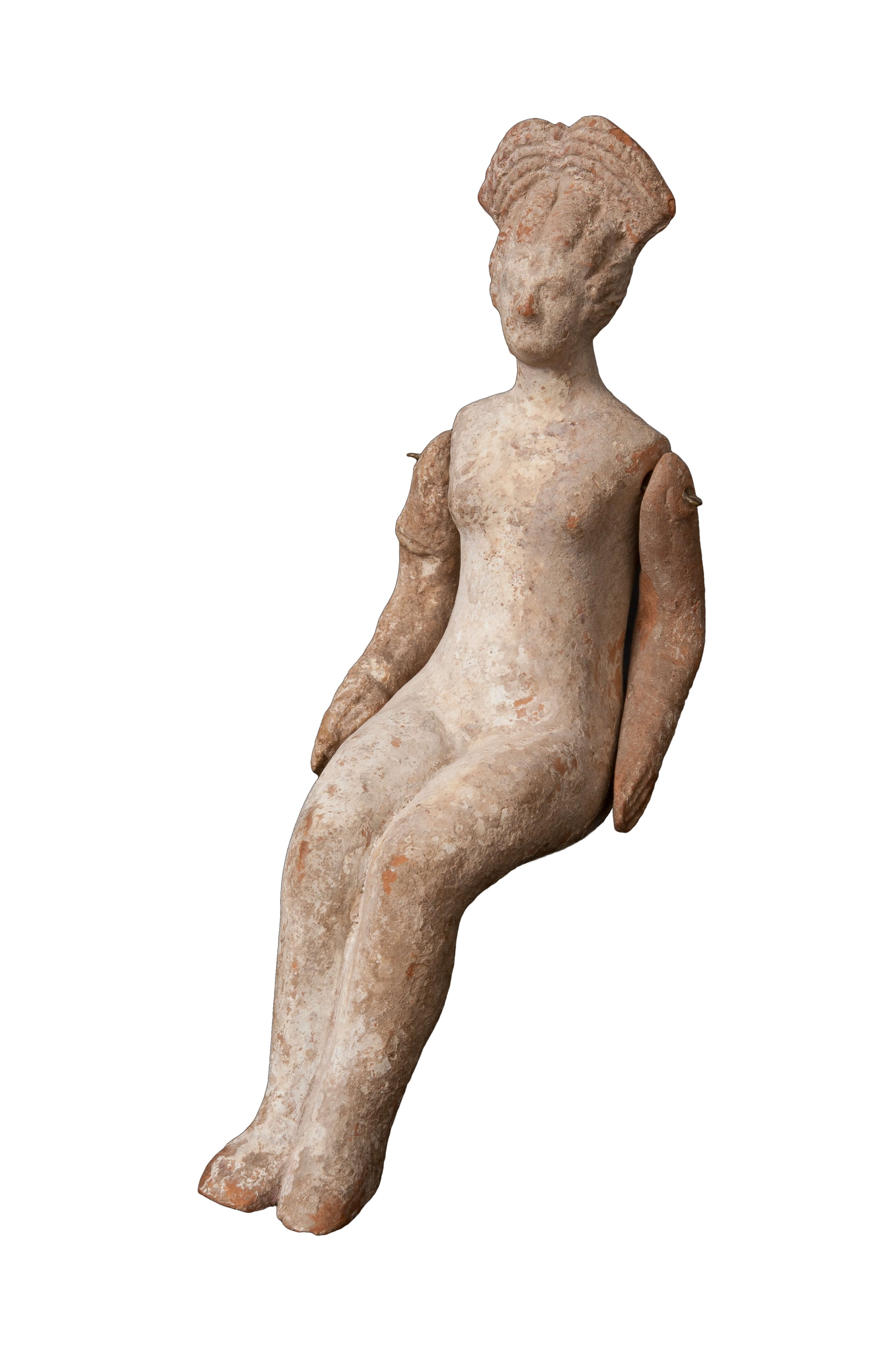 A terracotta doll in a simple form of a sitting woman. Her arms are by her side and her hair is arranged on her head.