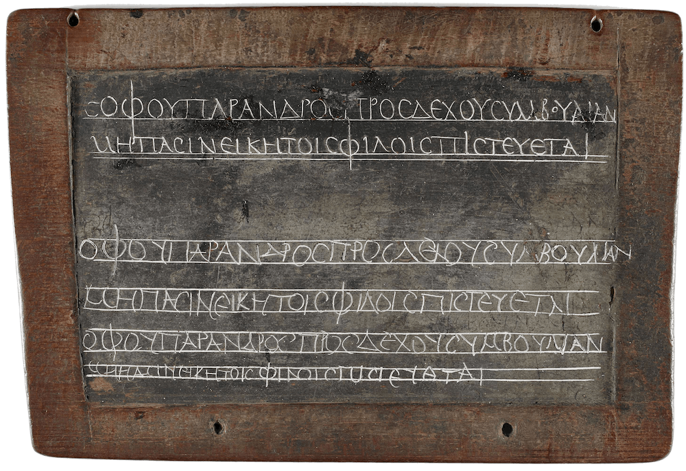 A rectangular wooden frame, the centre is filled with dark wax. On the surface Greek letters have been scratched on predrawn horizontal lines.