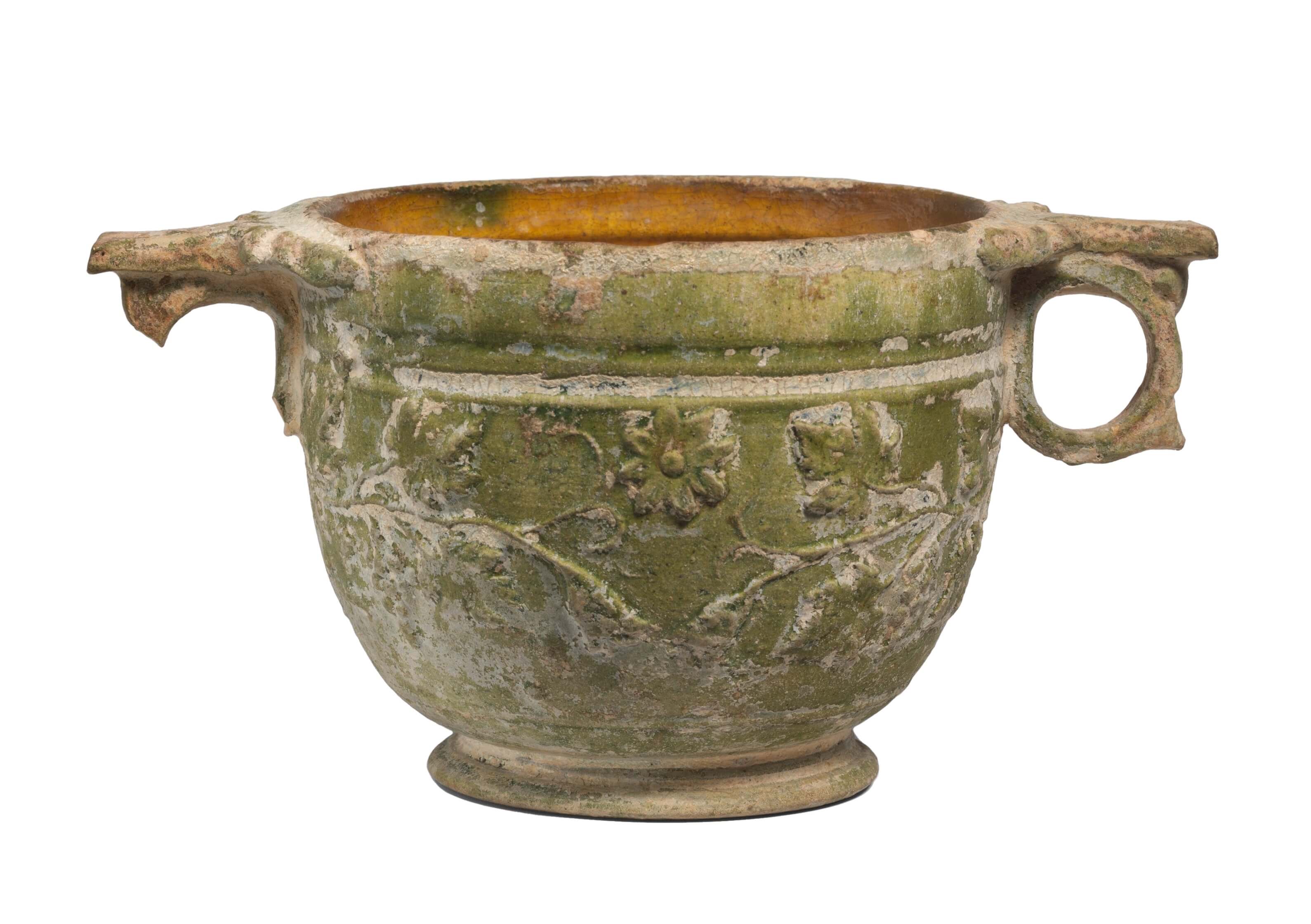 A small round cup with a circular handle on the right side. The matching handle on the left side has broken off. It is a green colour and has patterns of vines decorated on its surface.