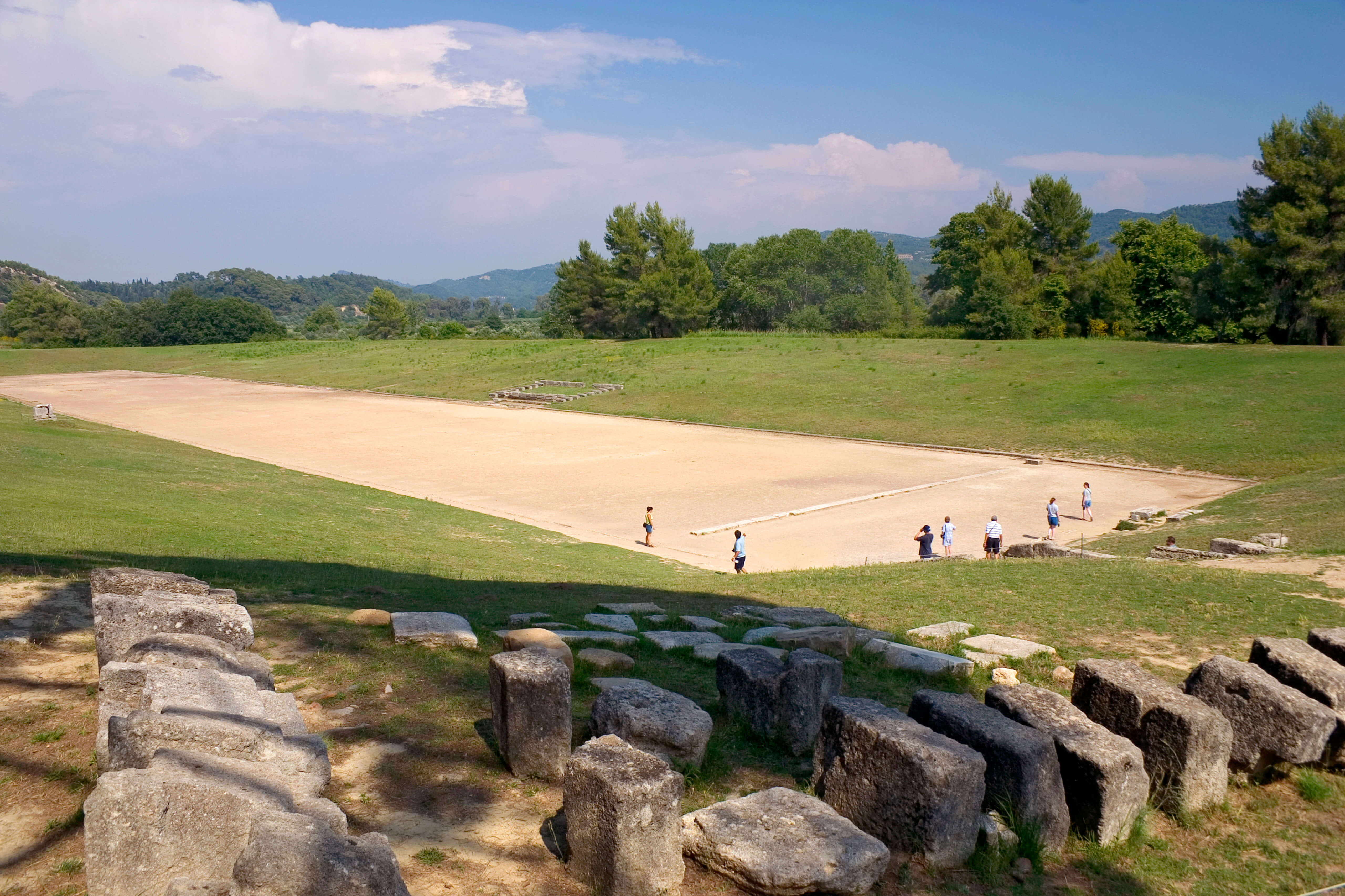 A photo of a long rectangular sandy space surrounded by grass banks on all sides that slope downwards towards the space. Beyound the grassy banks are trees and in the foreground are regular shaped blocks of stone on the floor. A few people are standing on the track.