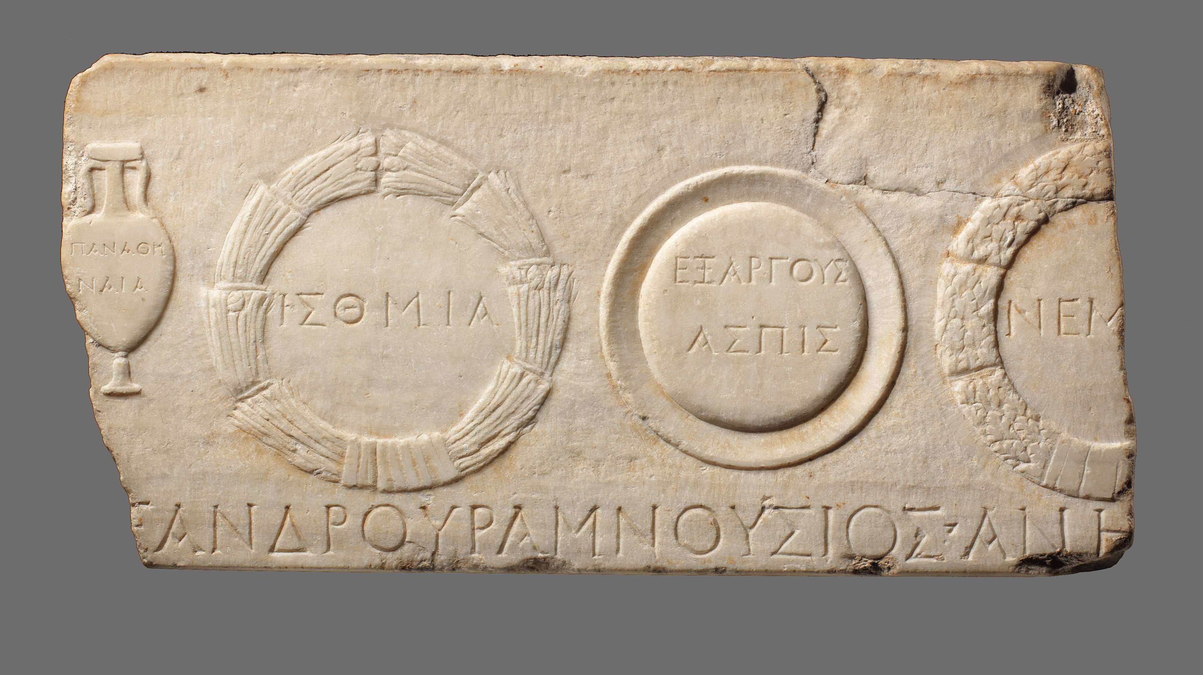 A rectangular piece of stone on which are carved circular wreaths and prizes and on the left and vase. Greek letters are written in the centre of the prizes and along the bottom.