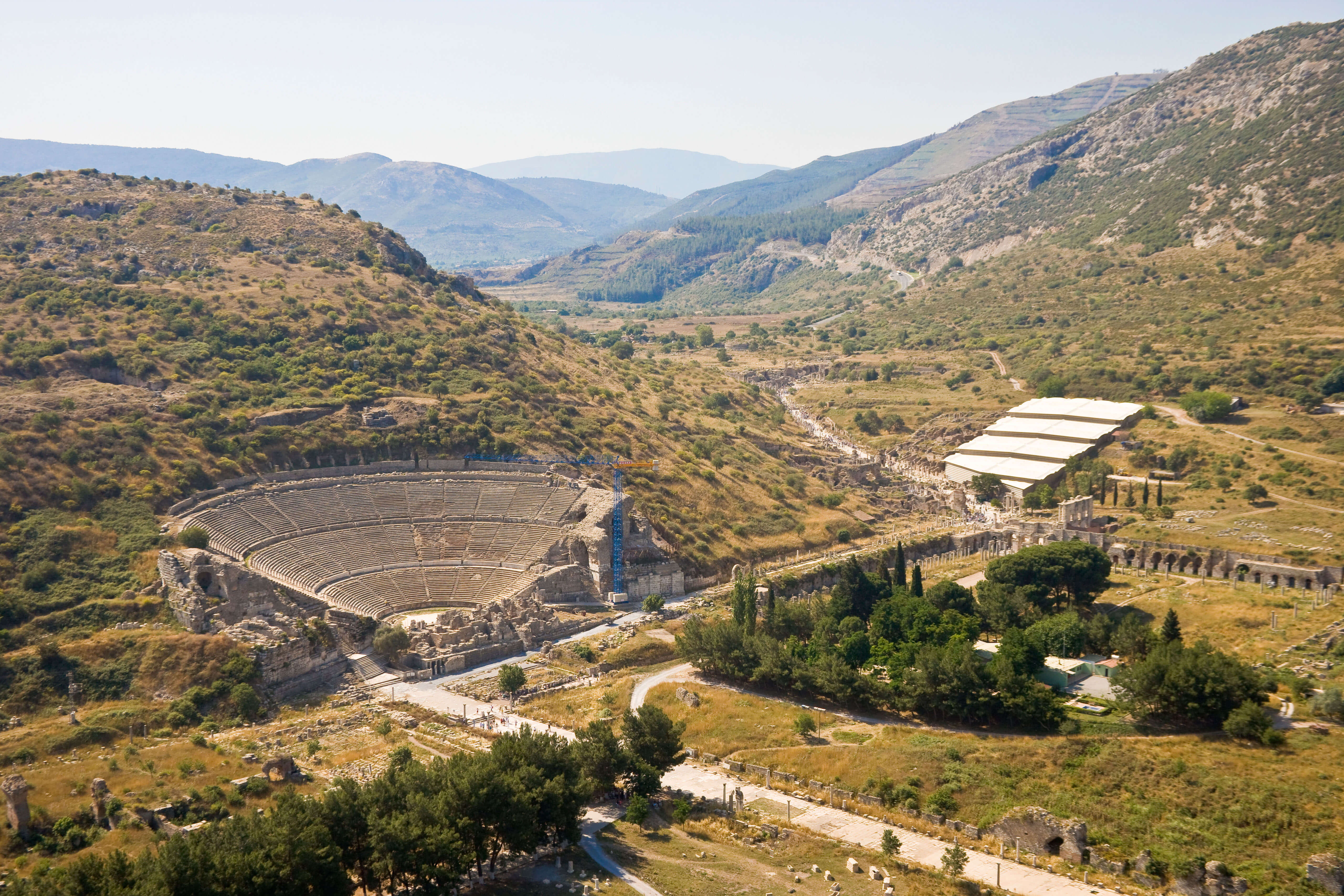 A photograph from above showing remains of a roman theatre and two big straight roads in a hilly, green landscape.