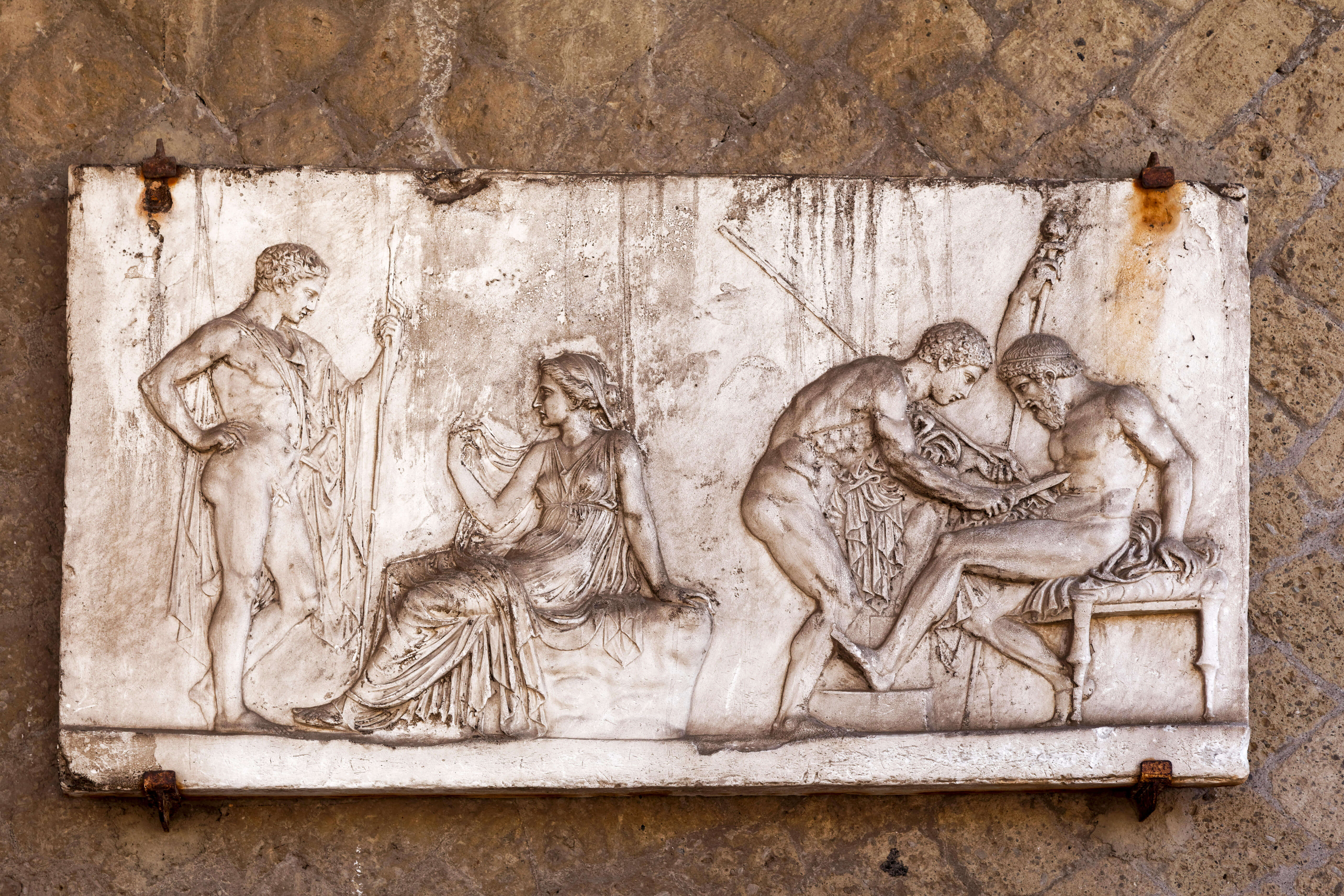 A stone relief showing a wounded man sitting on a stool with a spear in his abdomen. Another man is leaning over and extracting a spear using a knife in his right hand.