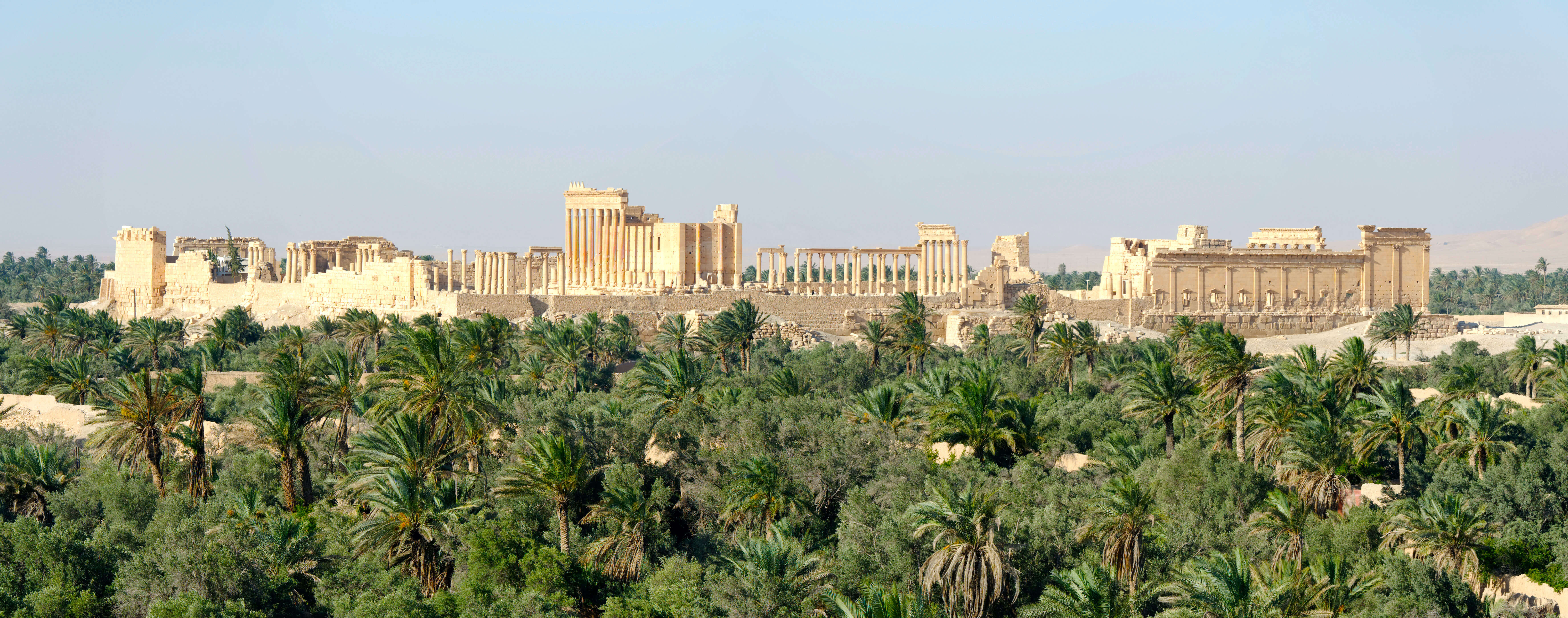 A panoramic photo showing the remains of a large temple complex. Many pillars and walls are still standing. The site is surrounded by a lush landscape of palm trees in the desert.
