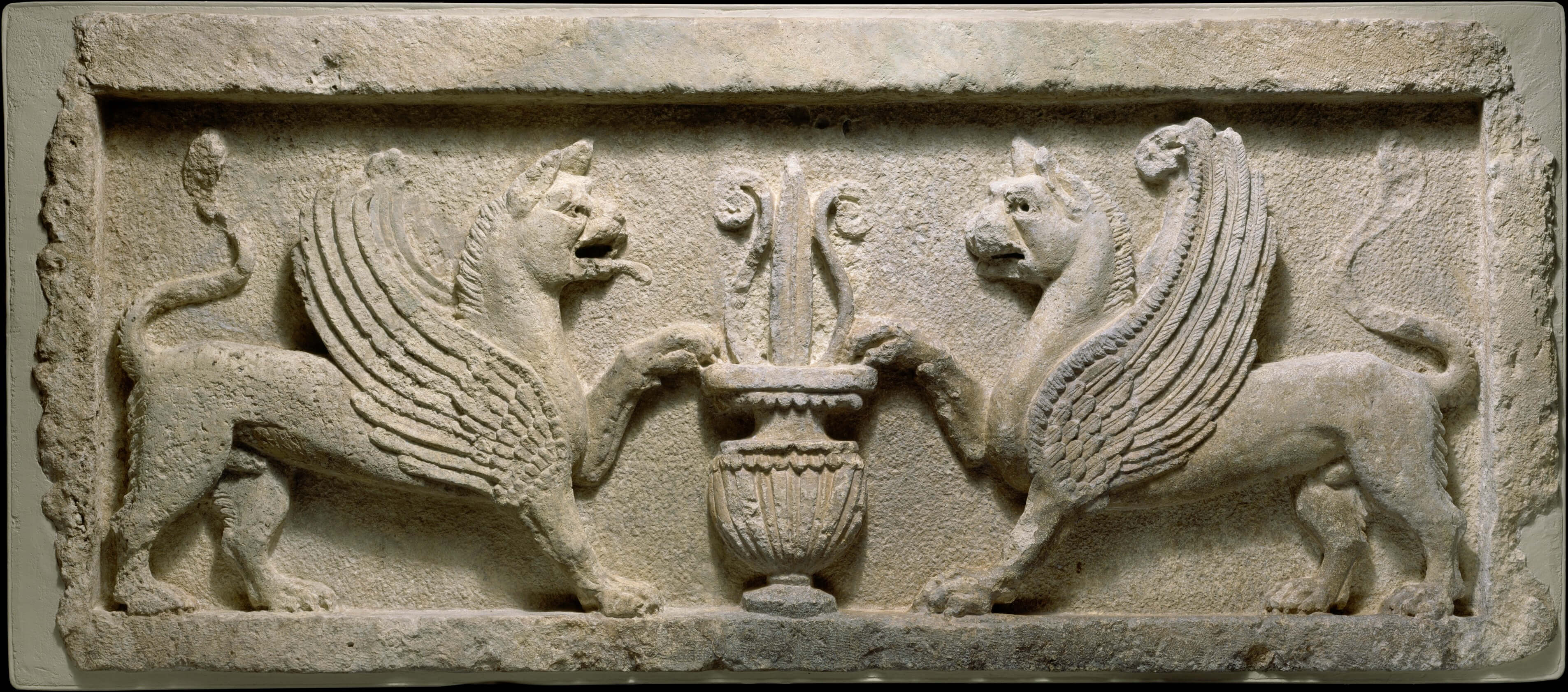 A stylized stone carving of two animals facing each other in a symmetrical design. They have the bodies of lions, but they both have wings. Between them is an ornately decorated vase.