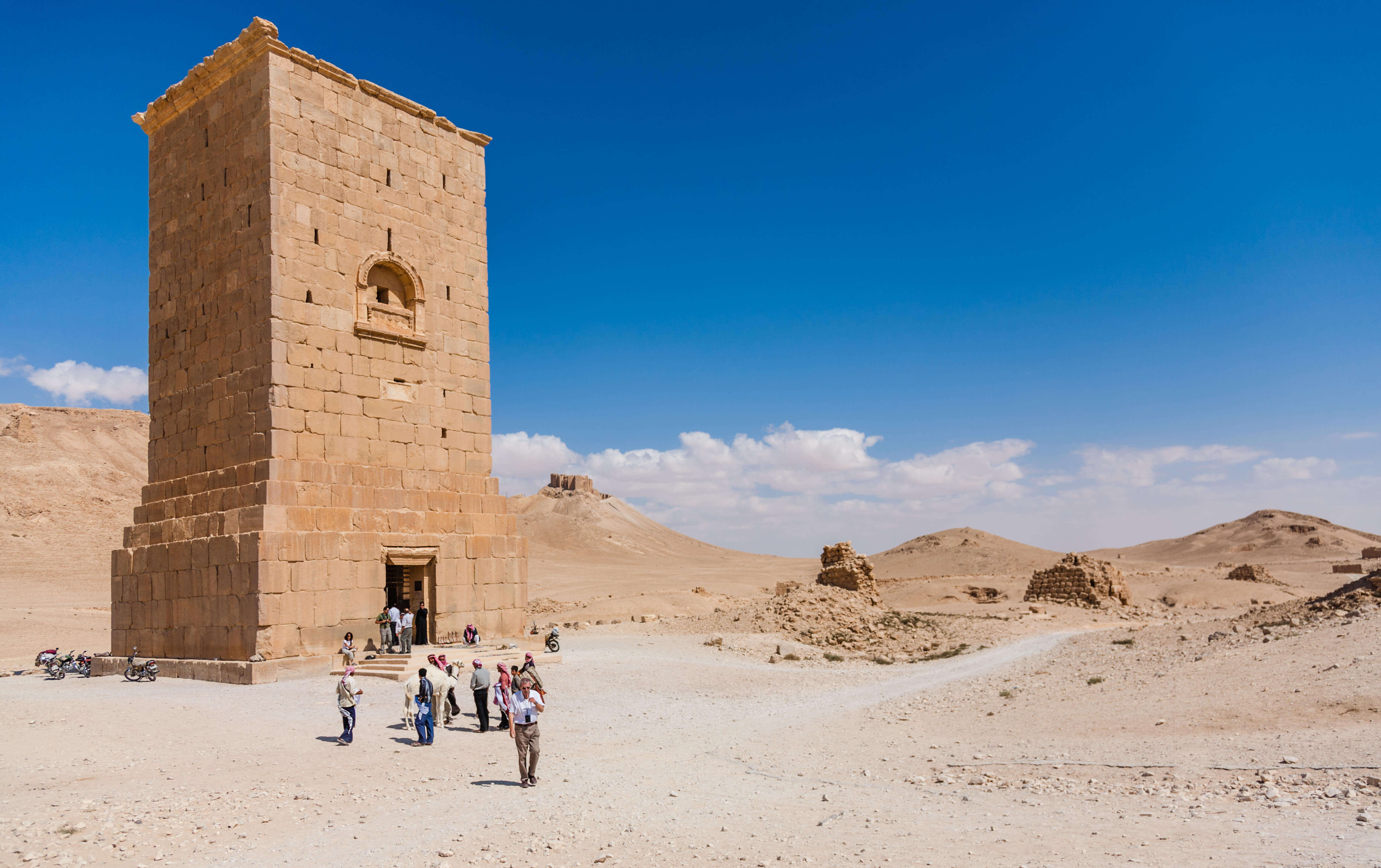 A rectangluar tower made of bricks. At the bottom there is a small rectangular door and in the centre above is an arched feature with a small window. It is set in a desert landscape and there are a small group of tourists in the foreground.
