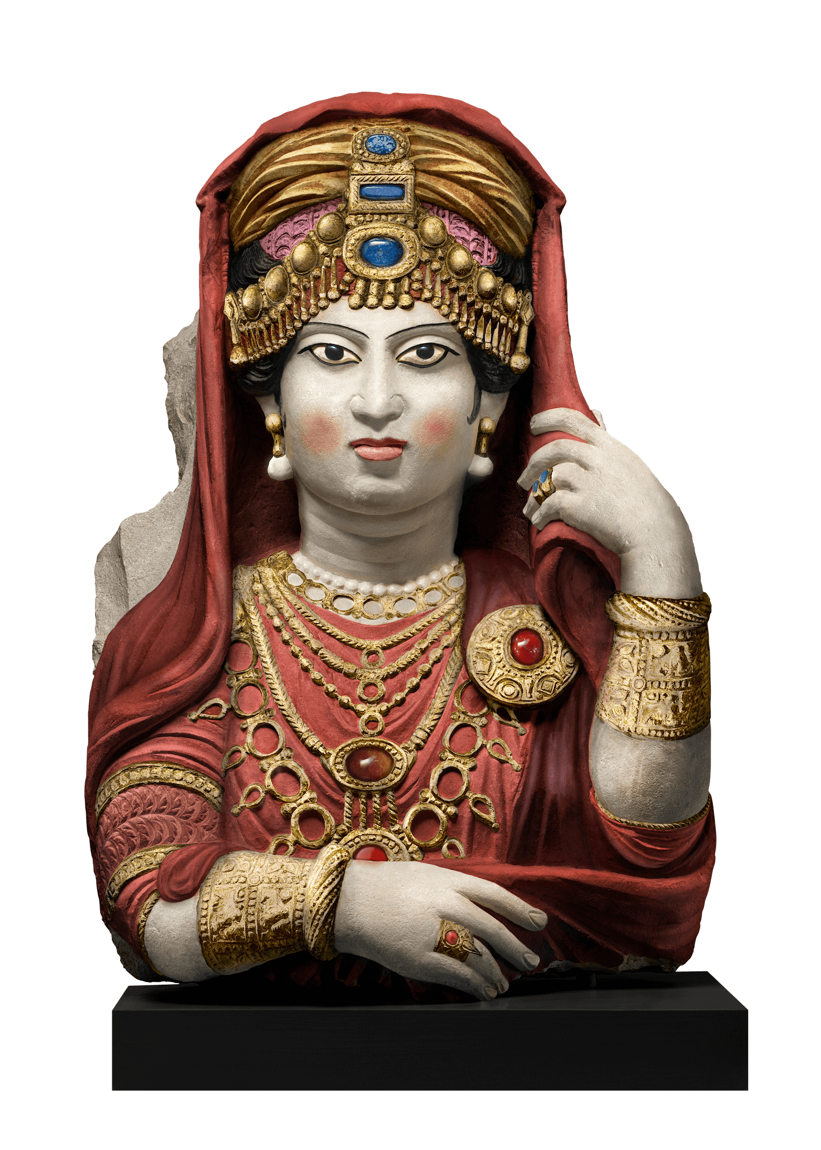 A statue of a woman with pale skin and dark rimmed eyes. She has an elaborate headress of red fabric with gold decorations. She wears a red dress and multiple gold neckalces, pearl earrings and gold armbands. Her left hand is reaching up to touch her headress.