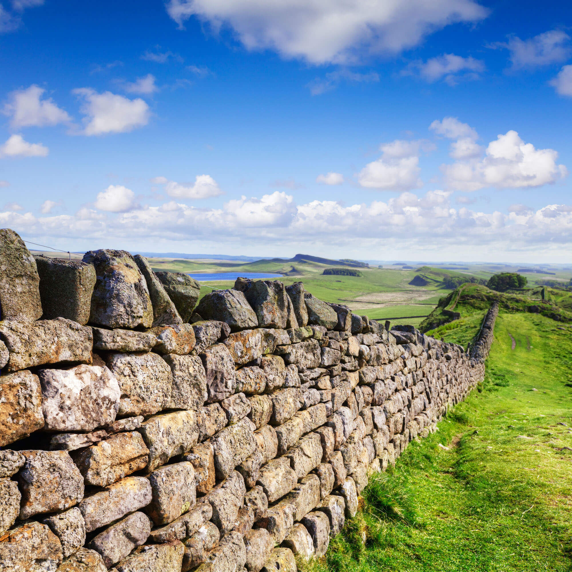 A panoramic photo showing a long stone wall crossing a green landscape of hills and strecthing into the distance. The stones are large and fitted together without cement.