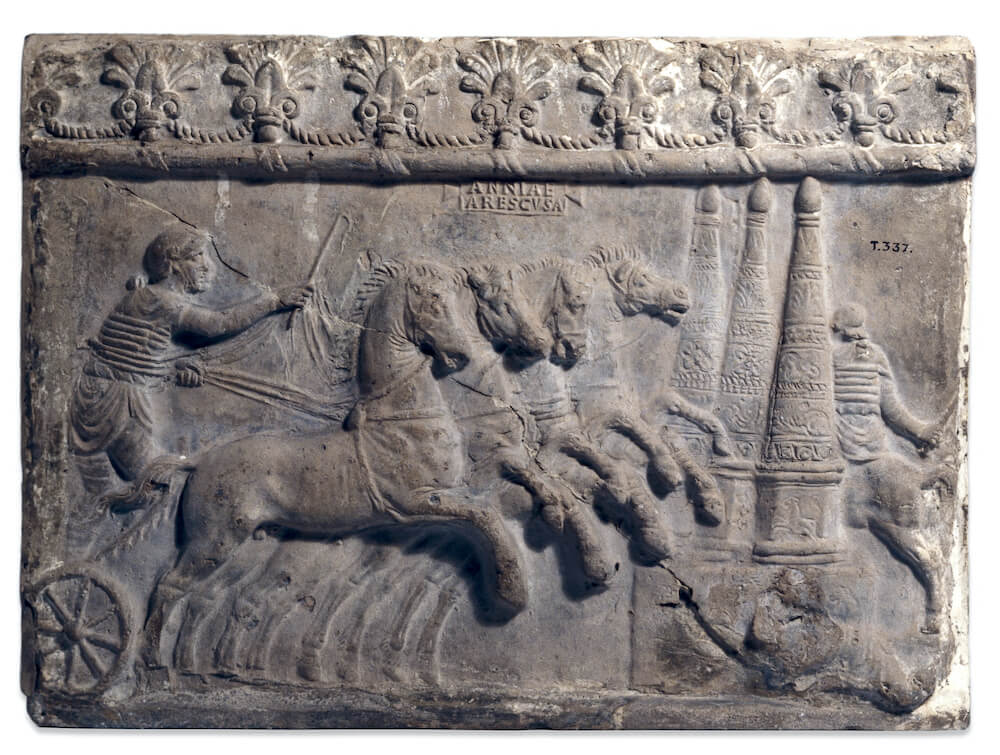 A rectangular clay tile decorated with a relief of a four horse chariot driving from the left. The charioteer holds the whip above the horses. On the right are the three conical metae.