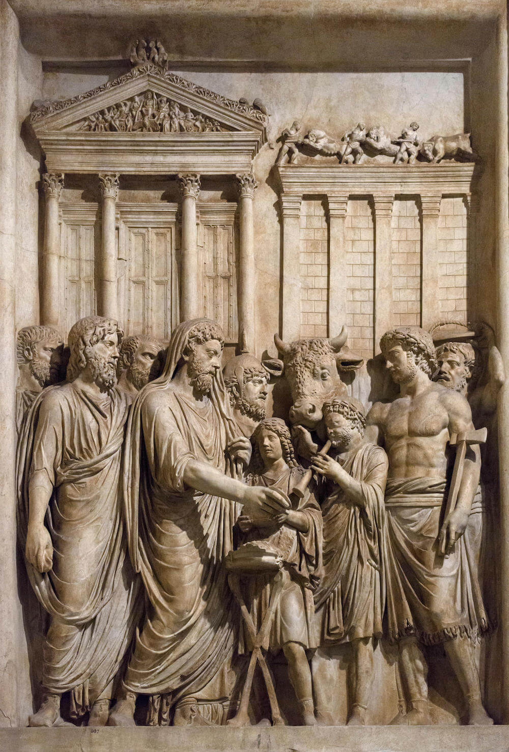 A stone carving showing men in togas standing at the front of a temple. In the centre is an ox and on the right is a man carrying an axe.