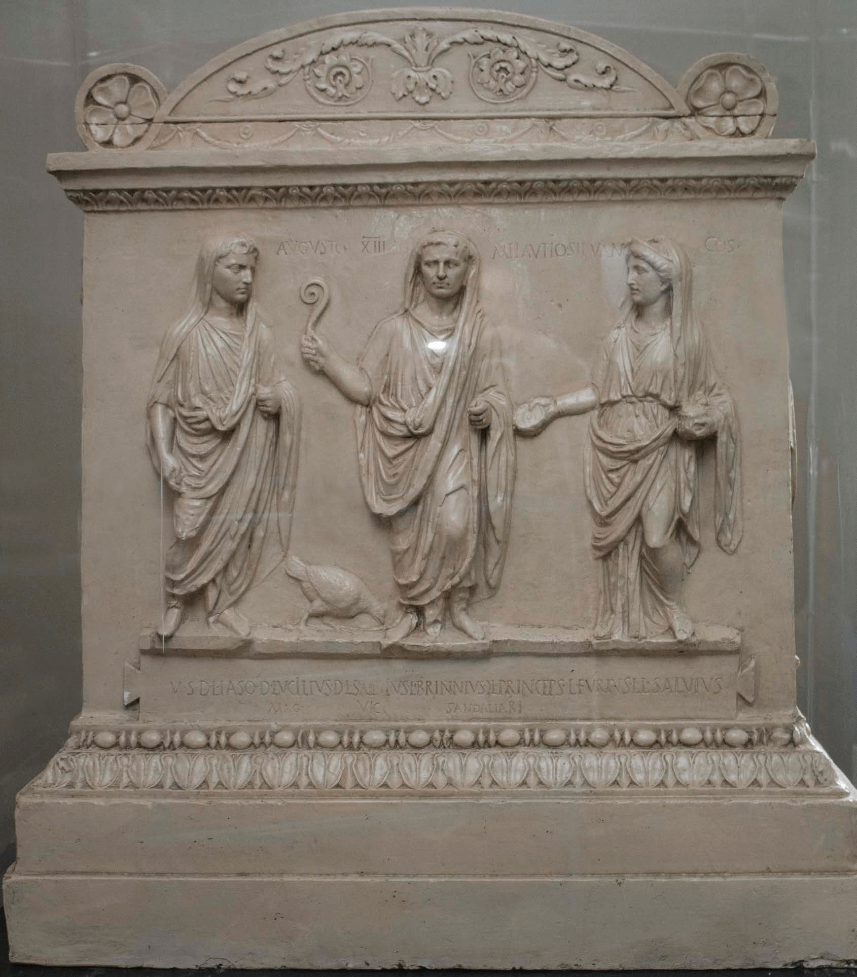 A rectangular stone altar with a curved top the borders of which are decrated intricately with patterns and leaves and flowers. In the centre three men stand wearing togas which have been pulled up over their heads. The man in the middle holds a stick with a curled end and at his feet is a chicken.