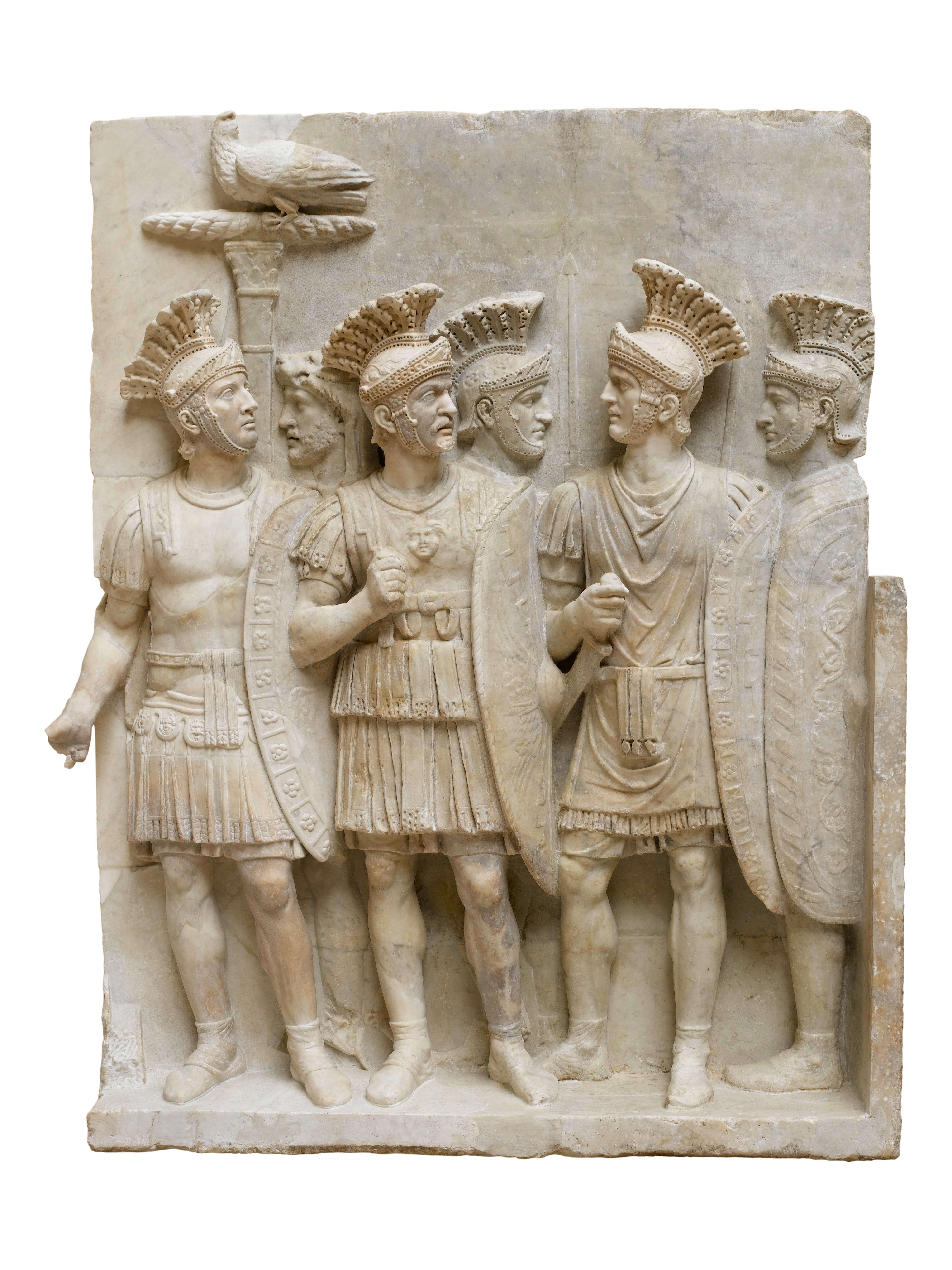 A stone relief showing a group of six soldiers standing in full armour holding shields. They have helmets with tall crests running front to back. One holds a pole, on top of which sits an eagle with his wings folded.