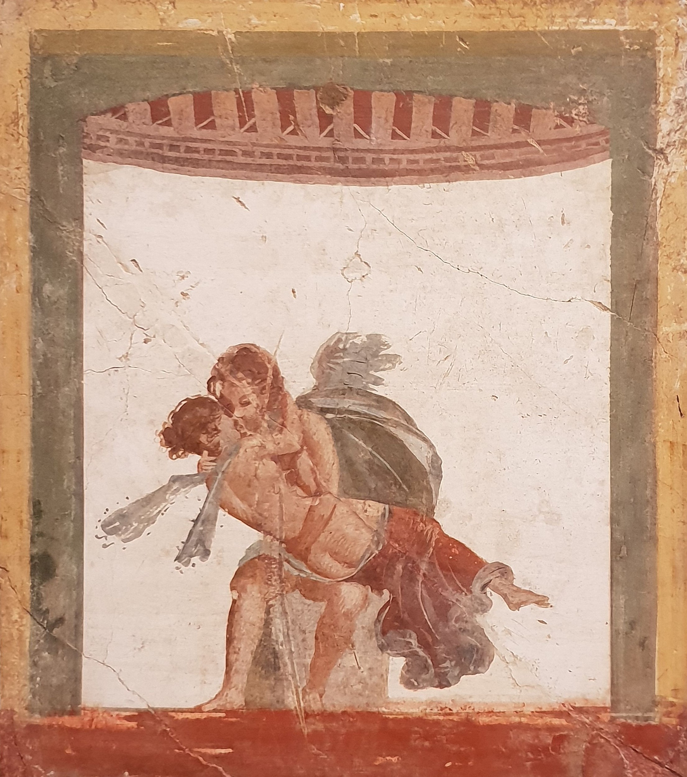 A winged man with a beard kisses a winged woman who is sitting on his knee. Around them is a decorative arch in reds and greens.